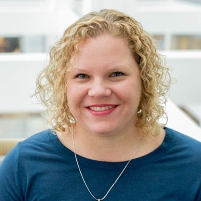 The image shows a woman with blonde, curly hair smiling at the camera. She has fair skin and is wearing a blue shirt and a silver necklace. The background appears to be a bright, modern indoor space, possibly an office or public building, with blurred architectural details.